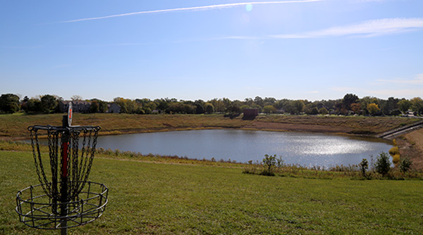 A disc golf basket in the foreground with a pond in the background at Margreth Riemer Park